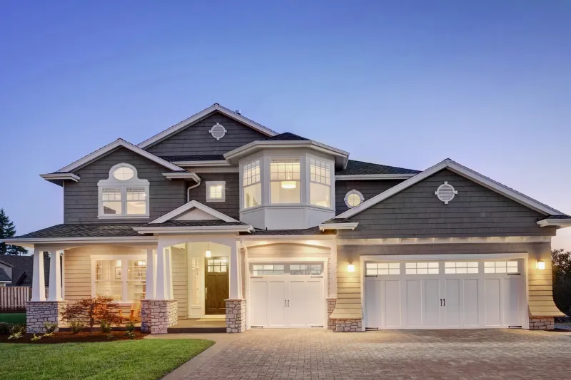 Two-story suburban house at dusk with gray siding, white trim, large front porch, three-car garage, and illuminated windows. The driveway and small landscaped yard are visible in front.