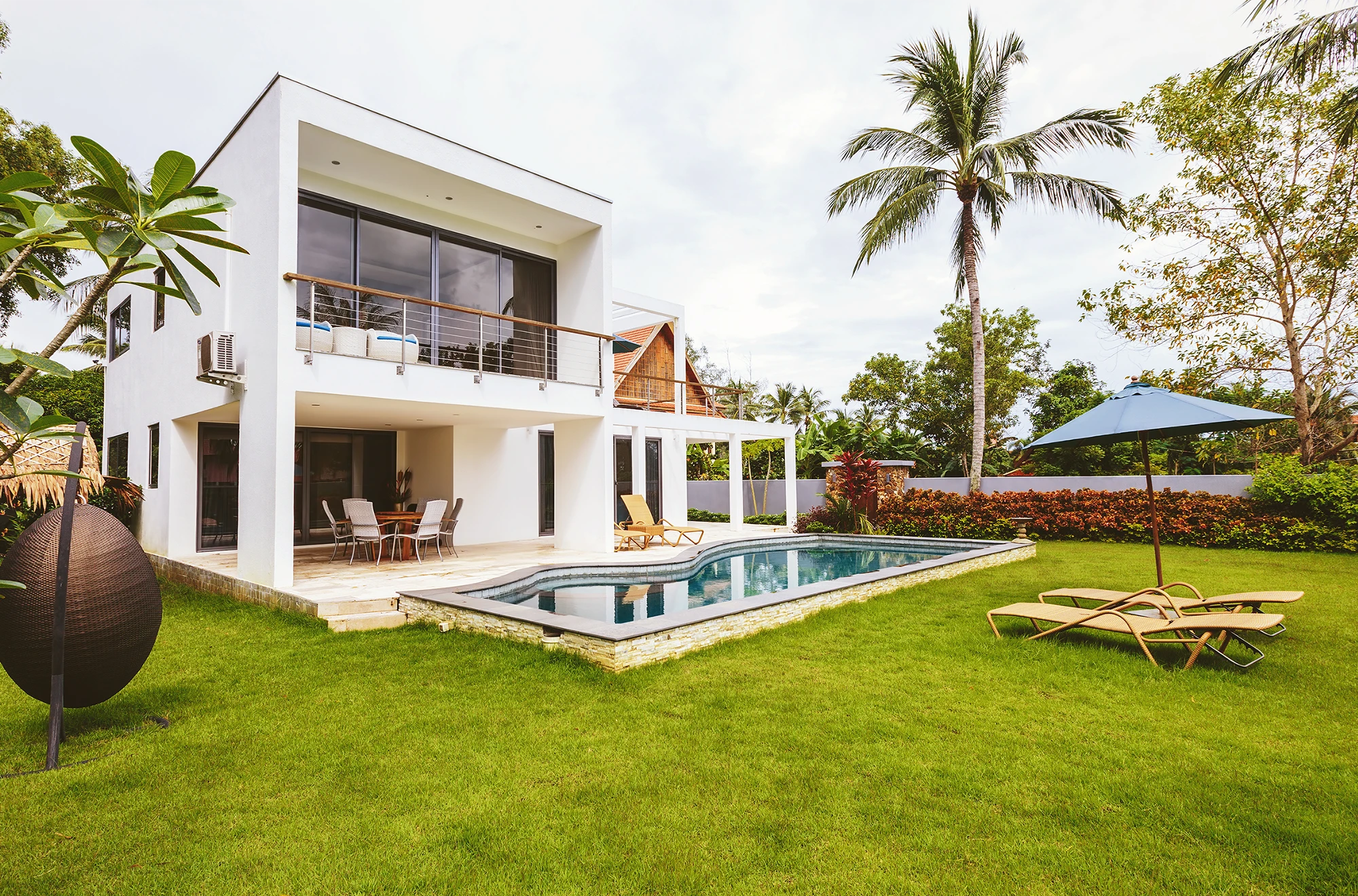 Modern white two-story house with large windows, a balcony, and a patio. There is a swimming pool, lounge chairs, a sun umbrella, tropical plants, and green lawn in the backyard. The setting appears tropical and inviting.