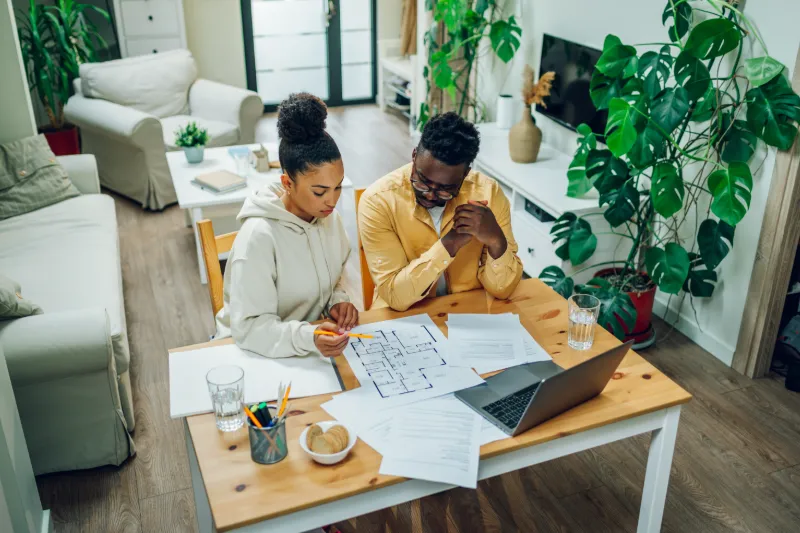 Two people sit at a table in a living room, looking at floor plans and papers. A laptop, pencils, water glasses, and documents to the Guide to the CalHFA MyHome Assistance Program are in front of them.